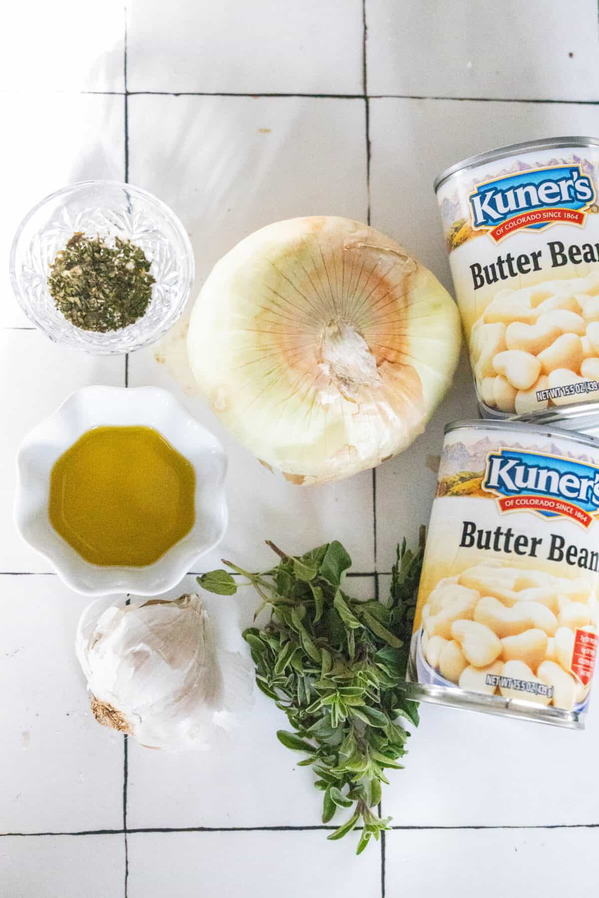 Cans of butter beans, an onion, garlic, and olive oil sit with dried oregano in a glass dish, accompanied by fresh green herbs on a tiled surface.