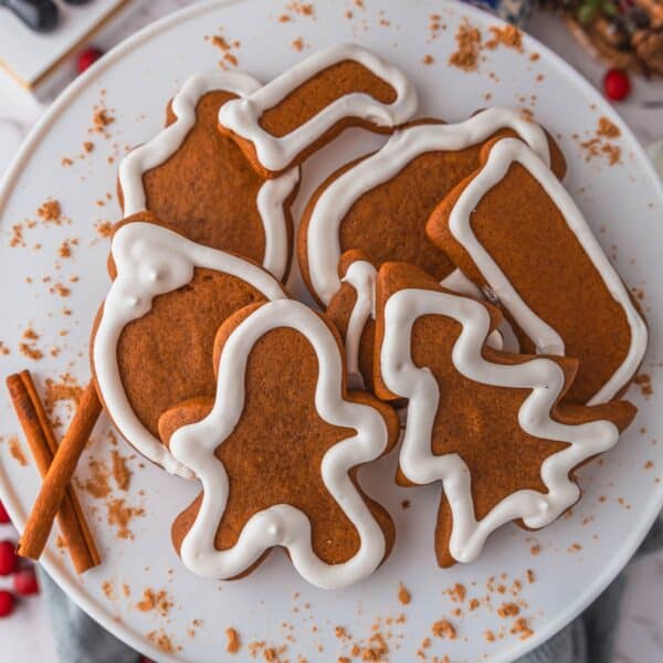 A plate of gingerbread cookies, artfully shaped into trees and bells, decorated with intricate white icing. Two cinnamon sticks rest beside the gingerbread cookies, adding a touch of aromatic spice.