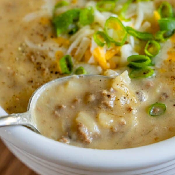 A close-up of a spoonful of Creamy Hash Brown Hamburger Soup reveals potato chunks, ground meat, green onions, black pepper, and shredded cheese, all nestled in a white bowl.