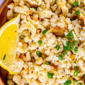 spaetzle noodles on a wooden plate with fresh pepper and sliced green onions on top next to a lemon wedge