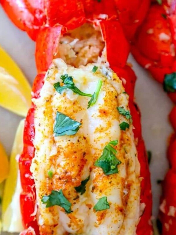 lobster tails seasoned with paprika and butter on a white plate with chopped parsley on top with lemons on the plate next to it