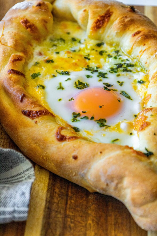 picture of baked khachapuri cheese bread on a wood cutting board with egg yolk and herbs on top 