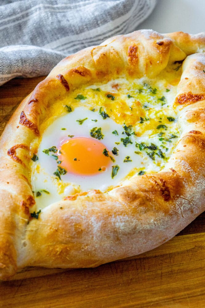 picture of baked khachapuri cheese bread on a wood cutting board with egg yolk and herbs on top 