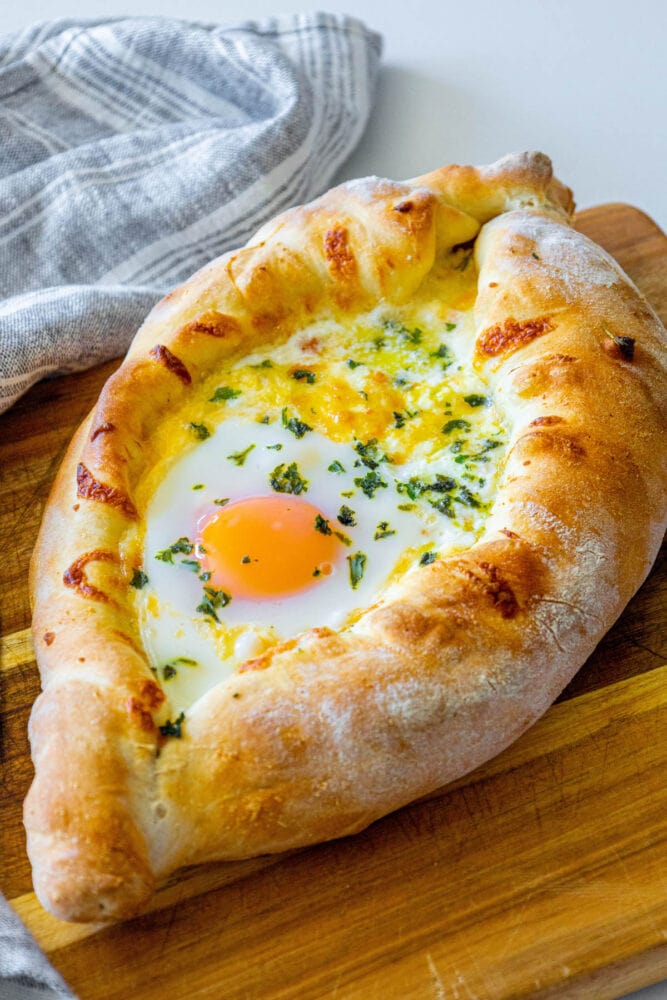 picture of baked khachapuri cheese bread on a wood cutting board with egg yolk and herbs on top 