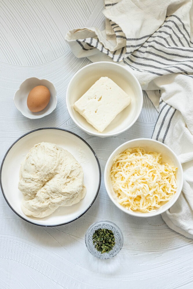 picture of dough, egg, feta cheese, shredded Muenster cheese, herbs on a table