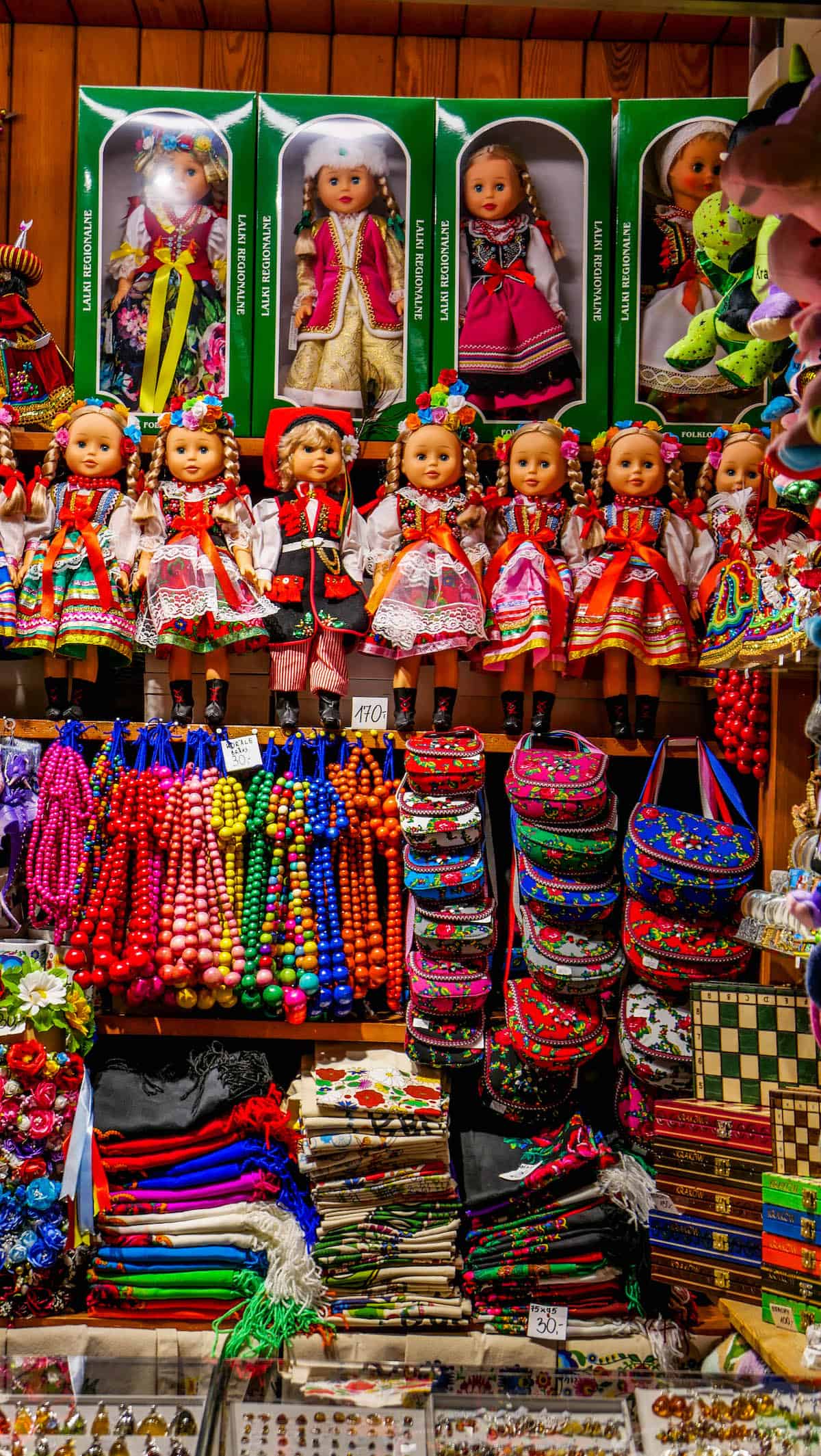 A display of dolls in traditional attire, colorful beaded necklaces, patterned bags, and assorted souvenirs reminiscent of the Krakow Christmas Market adorn the shelves.