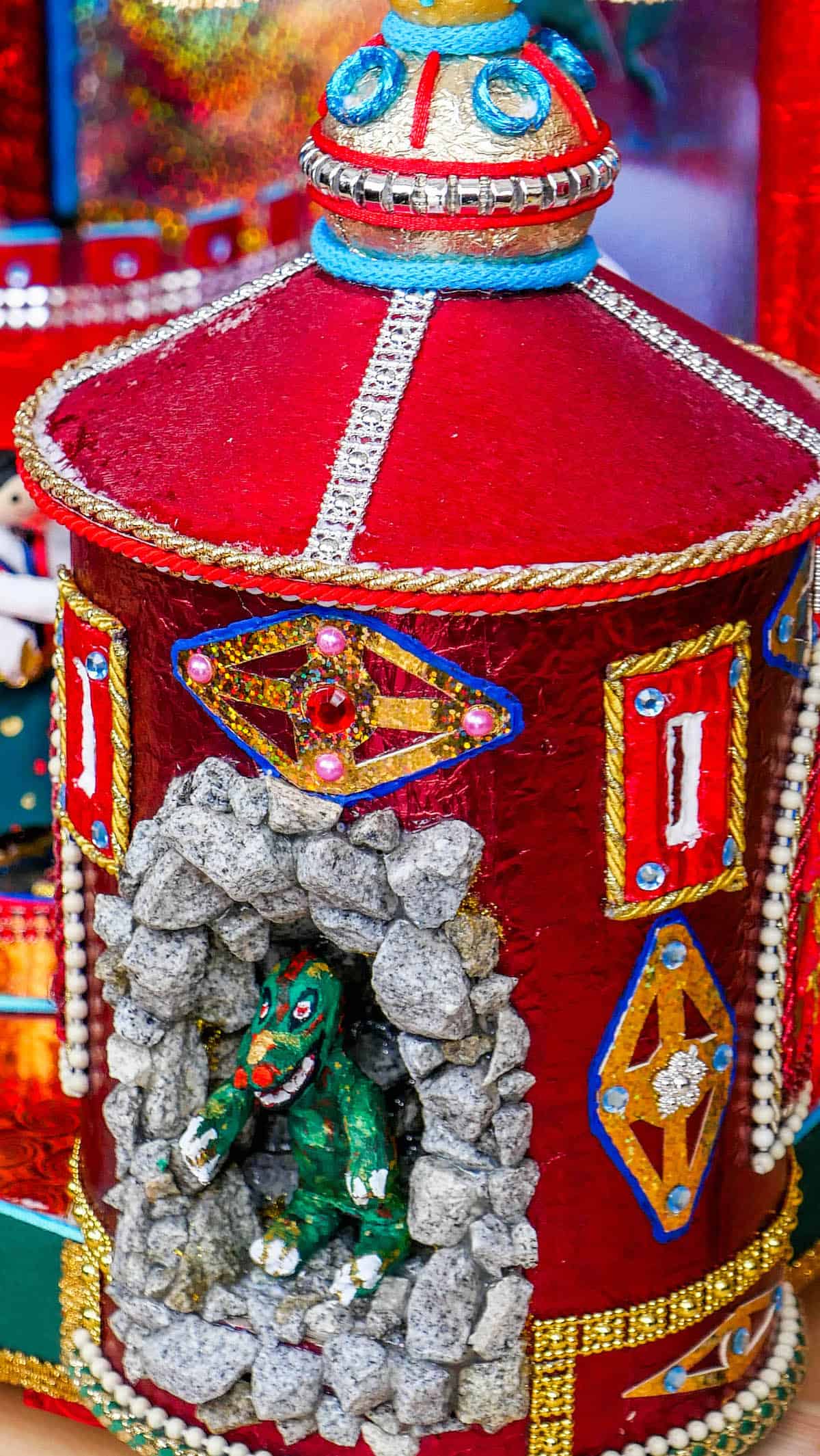 Colorful handcrafted decoration reminiscent of the Krakow Christmas Market, featuring a red structure adorned with rhinestones and a small green dragon figurine nestled in a stone-like alcove.