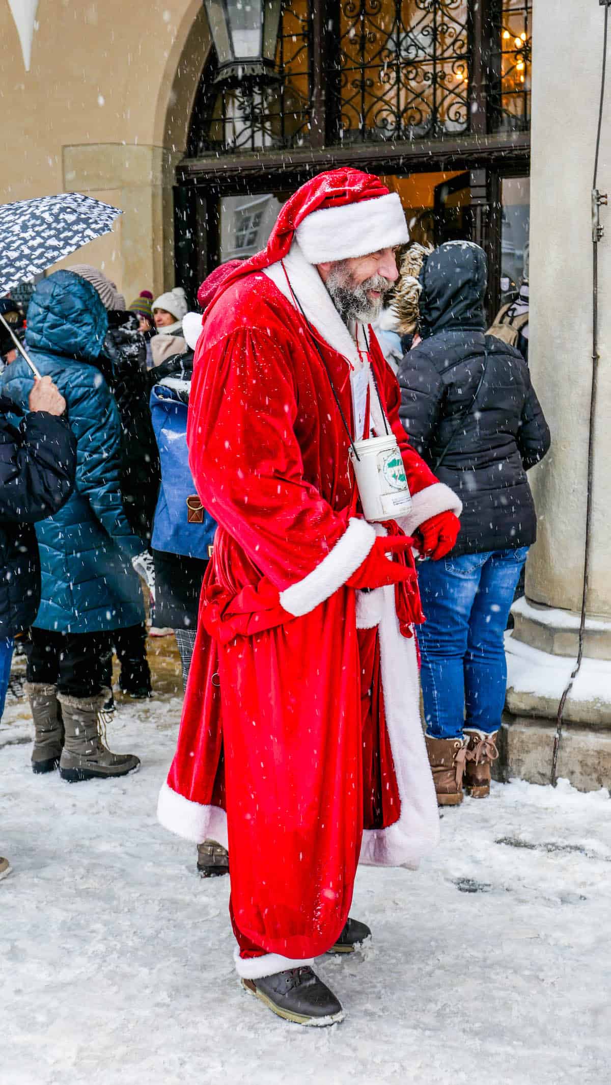 A man in a Santa costume stands joyfully amidst the snowy charm of the Krakow Christmas Market, surrounded by delighted holiday shoppers.