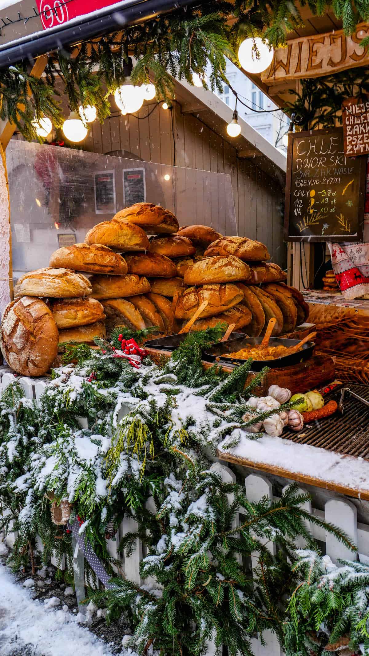 At the Krakow Christmas Market, a market stall boasts a large stack of bread loaves surrounded by snow-covered greenery and decorative lights.