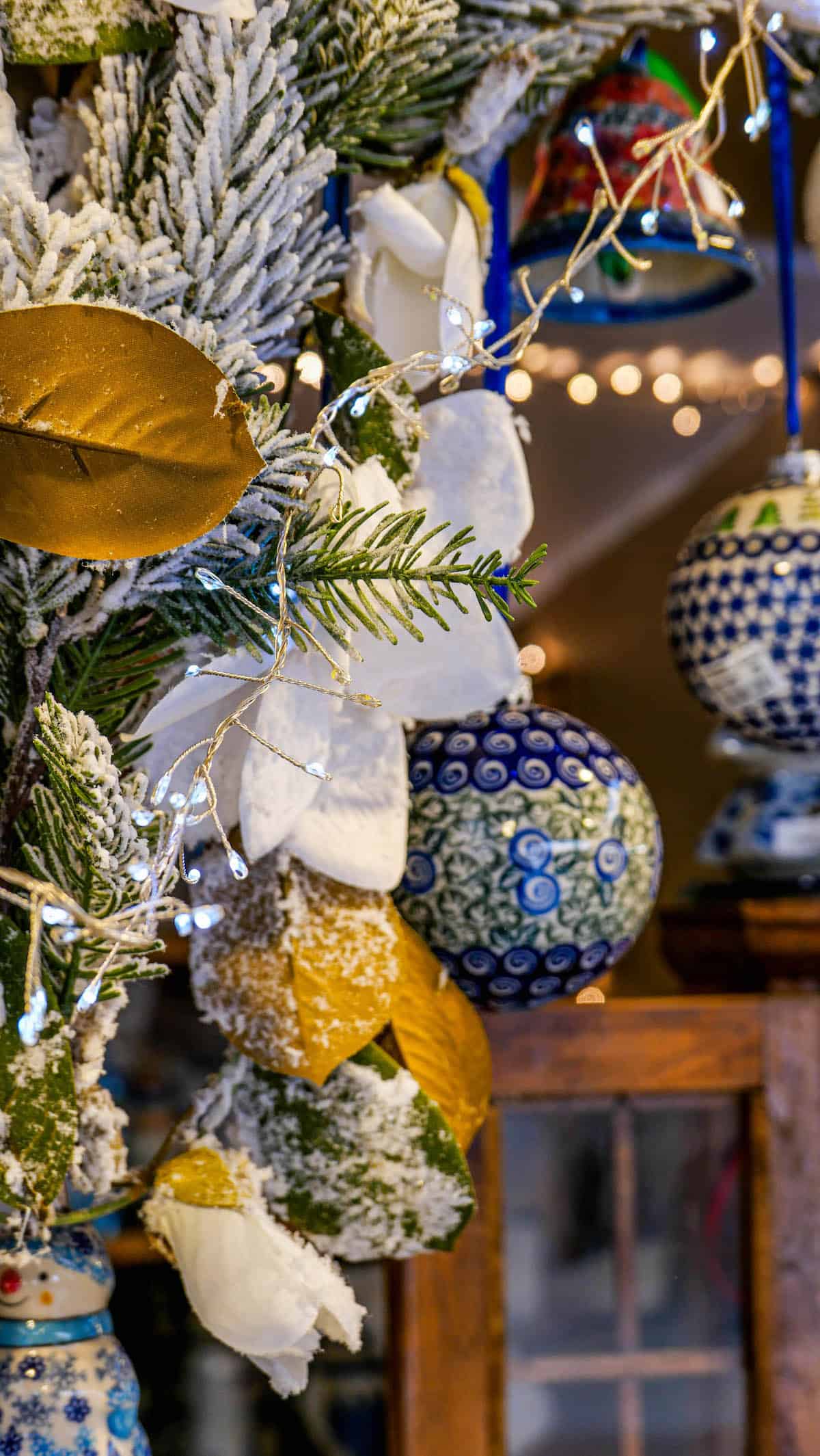 Festive decorations reminiscent of the Krakow Christmas Market feature blue and white patterned ornaments, snow-dusted foliage, and golden leaves hanging on display.