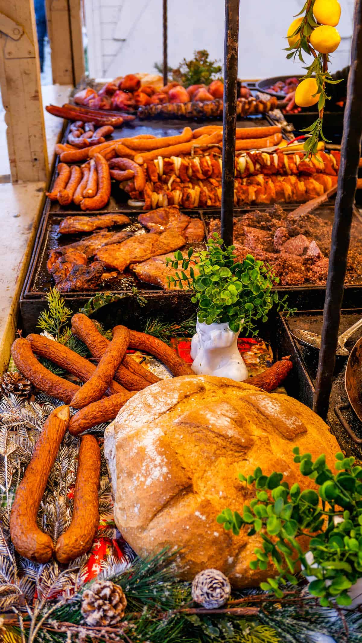 At the Krakow Christmas Market, an assortment of grilled meats, sausages, and skewers is artfully displayed alongside a large loaf of bread. Fresh herbs and decorative lemons add vibrant touches to this festive culinary scene.