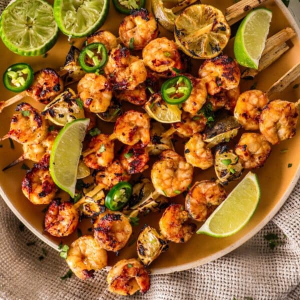 Margarita grilled shrimp skewers garnished with lime wedges, sliced green peppers, and charred lemon halves on a beige plate.