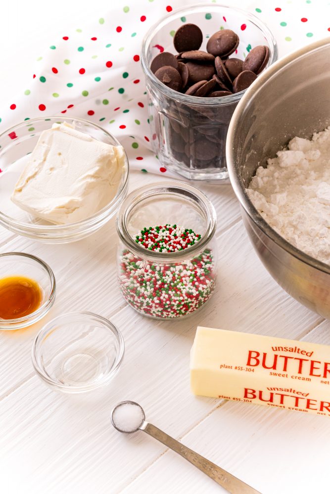 butter, a bowl of sprinkles, vanilla, cream cheese, chocolate, and powdered sugar in bowls on a table