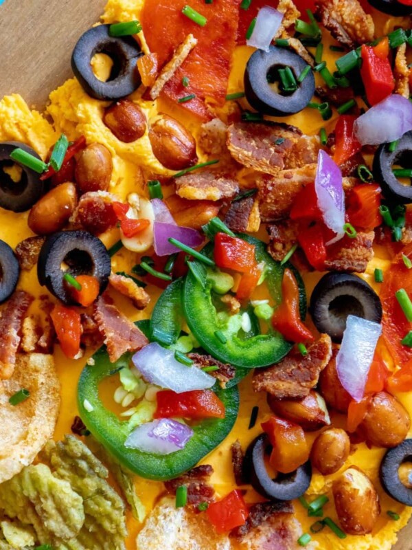 picture of pub cheese spread on a board covered with pork rinds, jalapenos, olives, bacon crumbles, peanuts, pimiento peppers, chives, red onion, and pepperoni