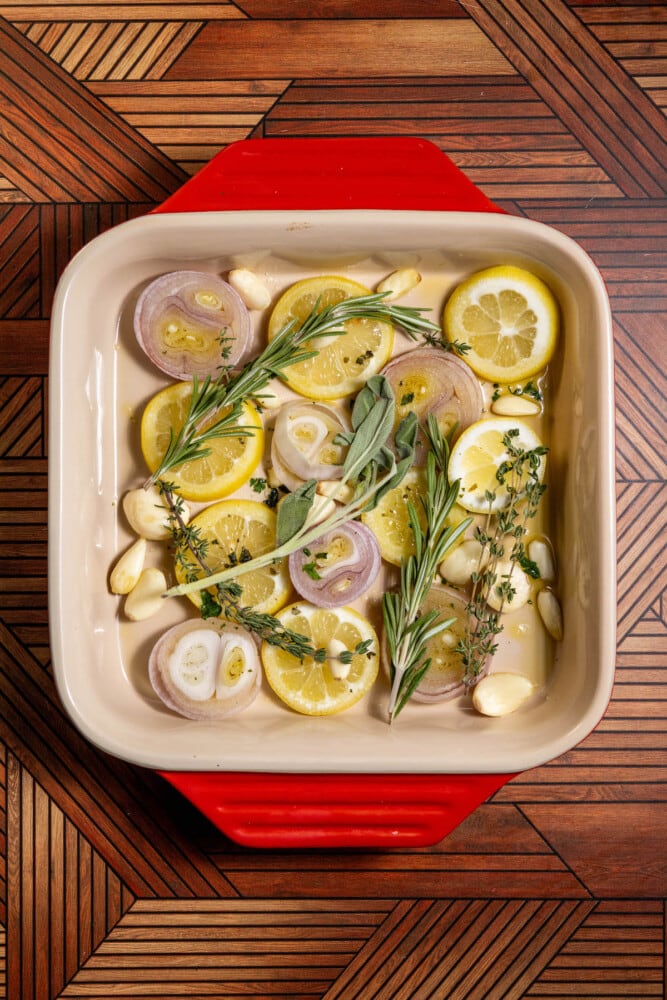 slices of lemon, slices of shallots, garlic cloves, and sprigs of herbs in a baking dish