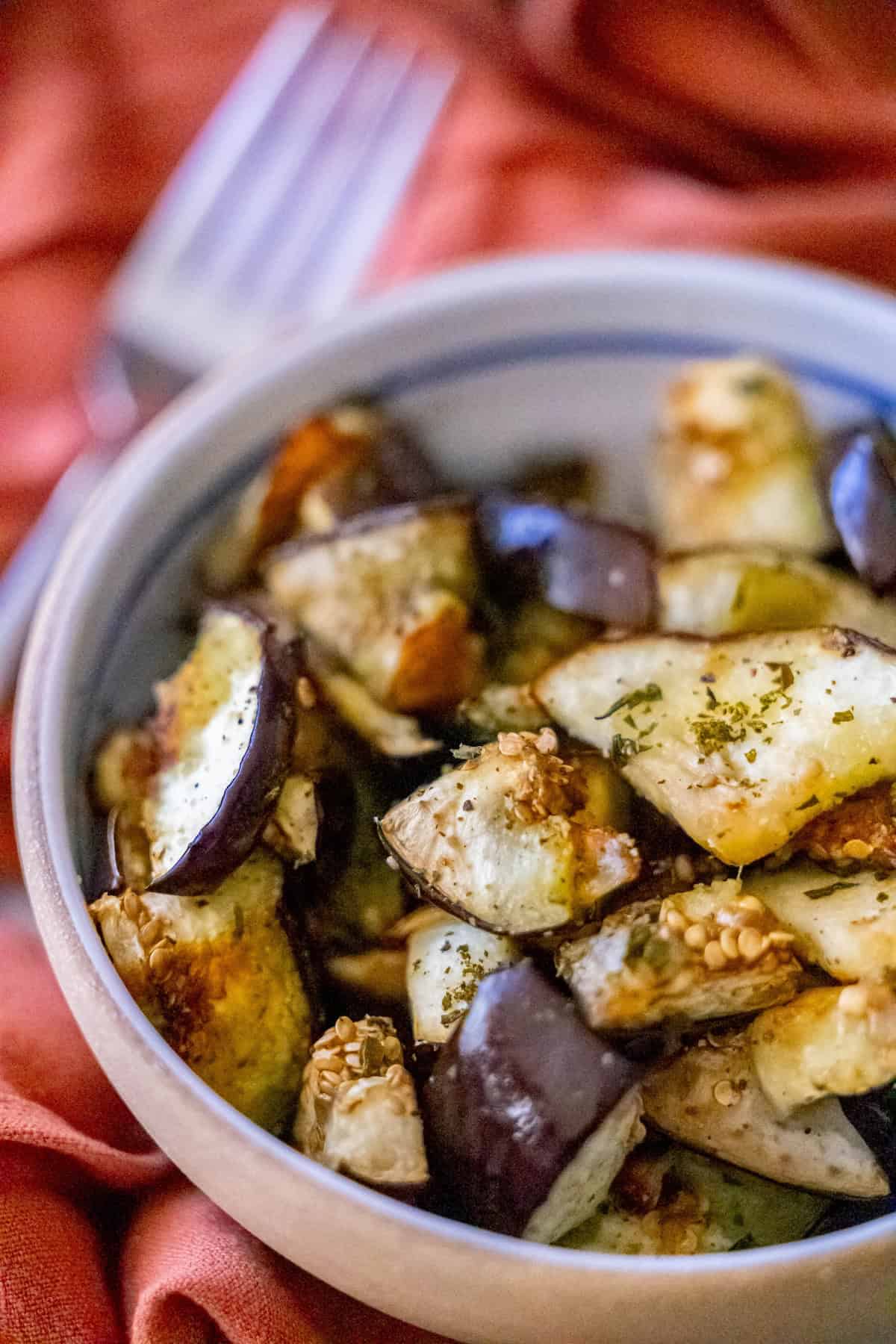 A vibrant bowl of roasted eggplant with fragrant herbs and sesame seeds sits on an orange cloth, exuding warmth. A fork waits in the background, inviting you to indulge in this delightful eggplant recipe, a perfect celebration of flavor and texture.