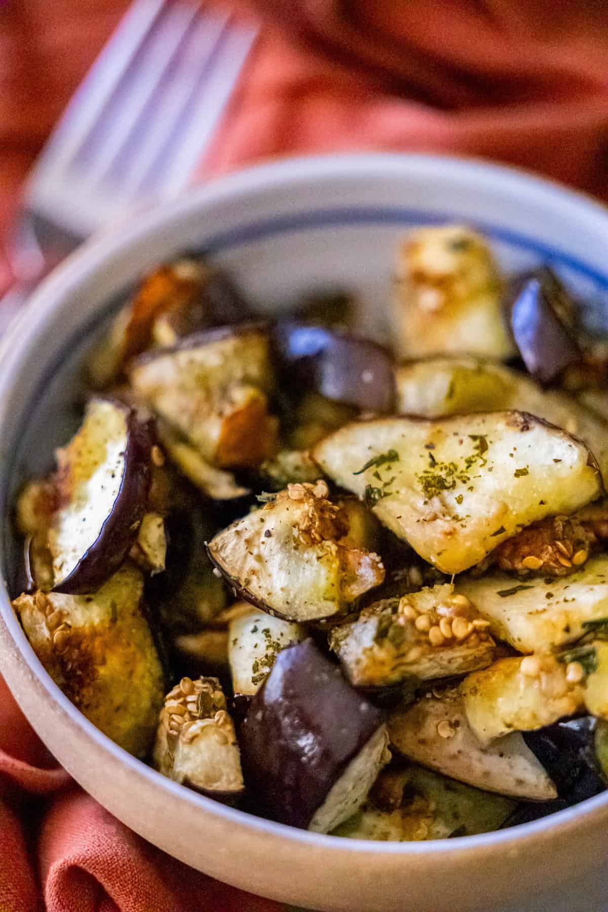 A bowl of roasted eggplant pieces, seasoned to perfection with aromatic herbs, rests on an orange cloth, while a fork lurks in the background. This delightful dish is a must-try for any eggplant recipe enthusiast.