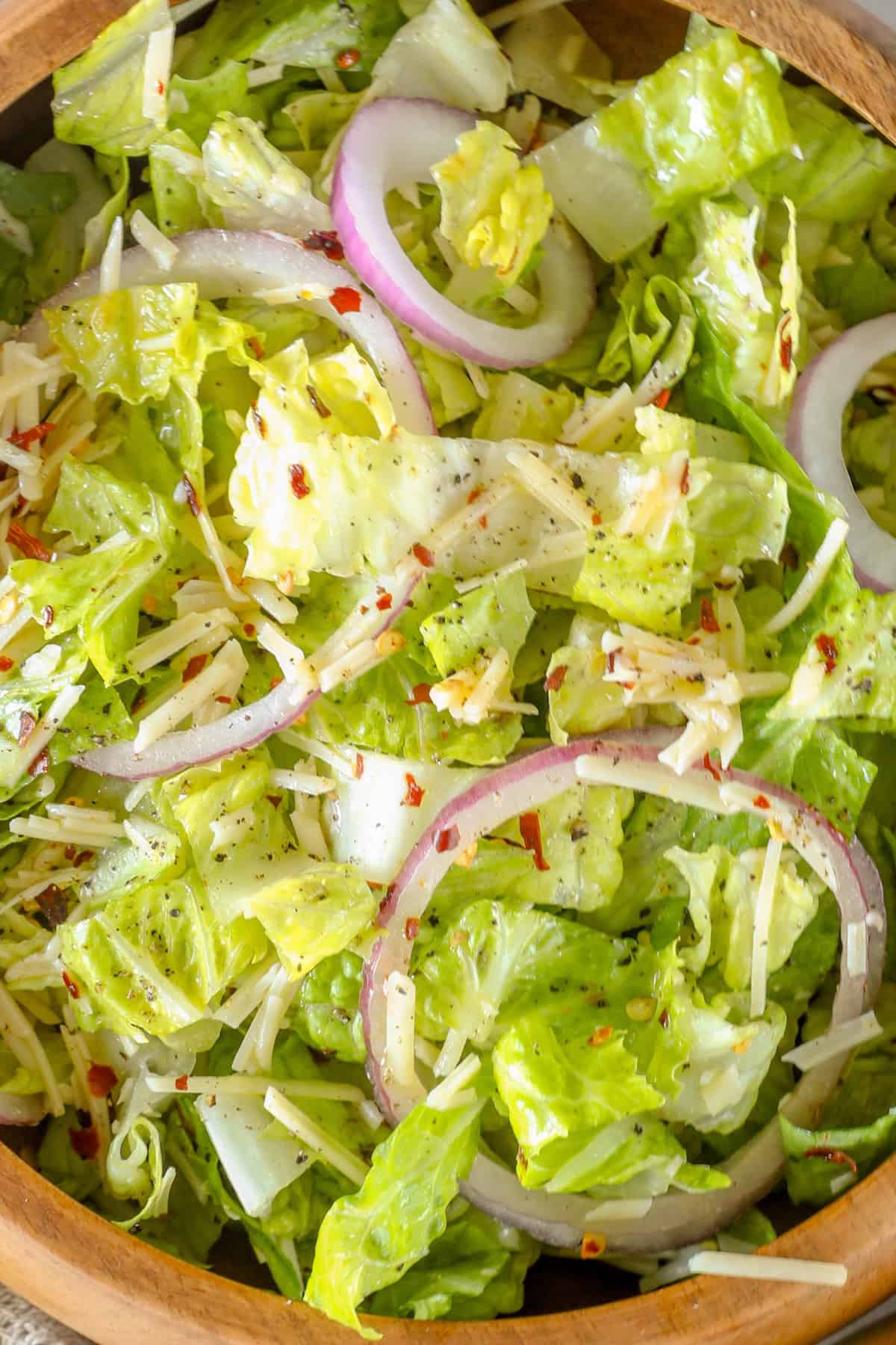 A wooden bowl filled with a vibrant romaine salad, featuring fresh lettuce, sliced red onions, grated parmesan cheese, and a sprinkle of red pepper flakes.