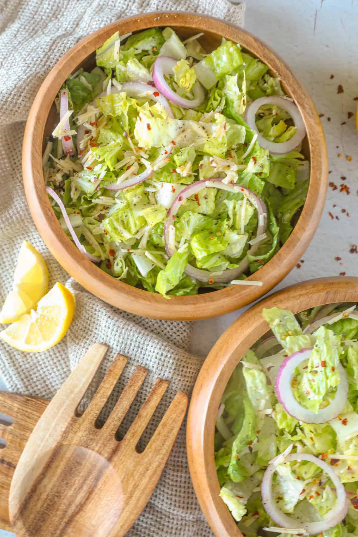 Two wooden bowls of fresh romaine salad with lettuce, sliced red onion, and a lemon wedge on a cloth, accompanied by wooden salad servers.