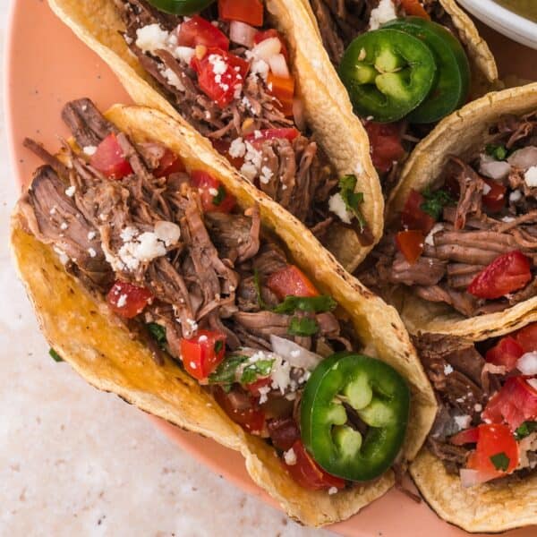 A plate of four shredded beef tacos topped with diced tomatoes, onions, jalapeños, and crumbled cheese.