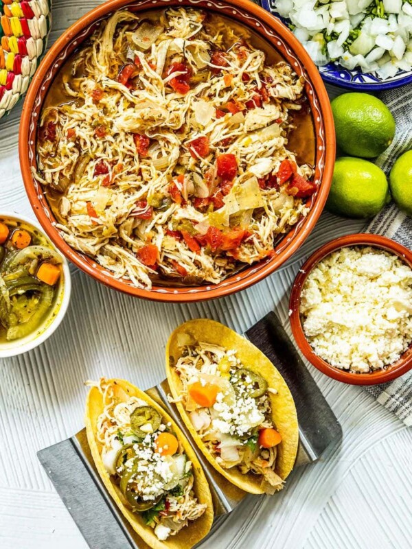 shredded chicken with tomatoes and green chile in a telavera bowl next to chicken tacos in a taco stand and a bowl of jalapeno and carrot salsa, a bowl of crumbled white cheese, some limes, a bowl of diced onions, and a bowl of tortillas on a table