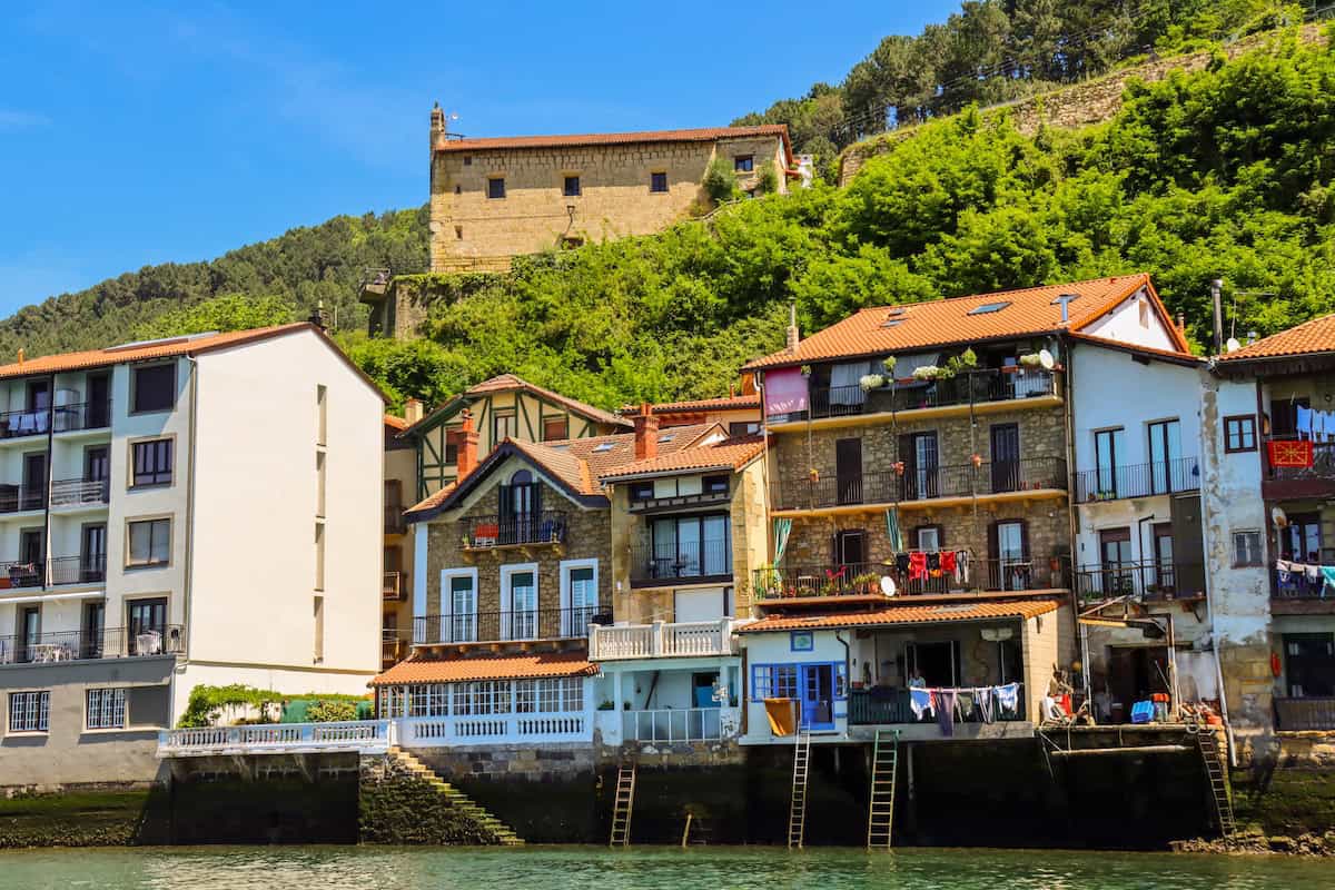 Colorful buildings line a waterfront below a hillside with greenery and a stone structure under a clear blue sky.