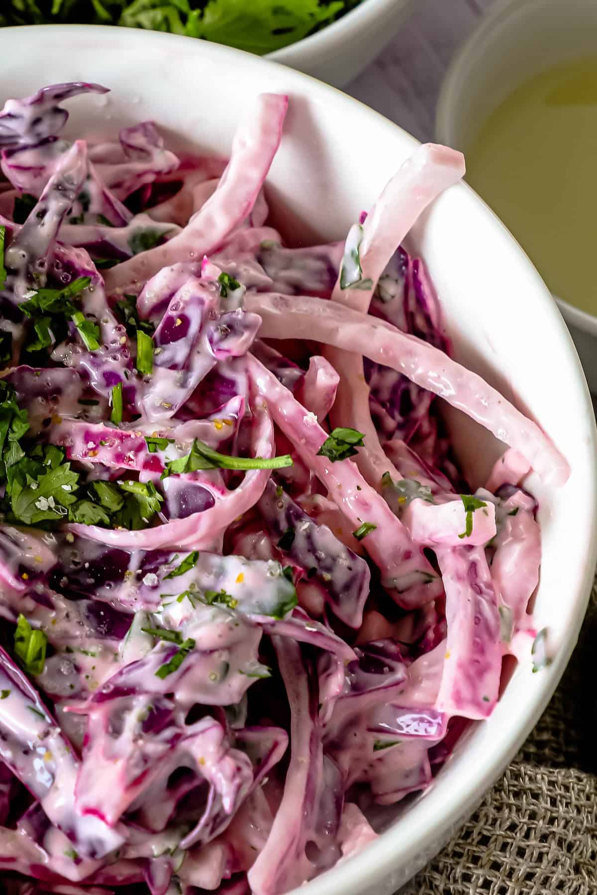 A close-up of a white bowl filled with vibrant purple coleslaw, garnished with chopped green herbs and mixed with a creamy dressing.