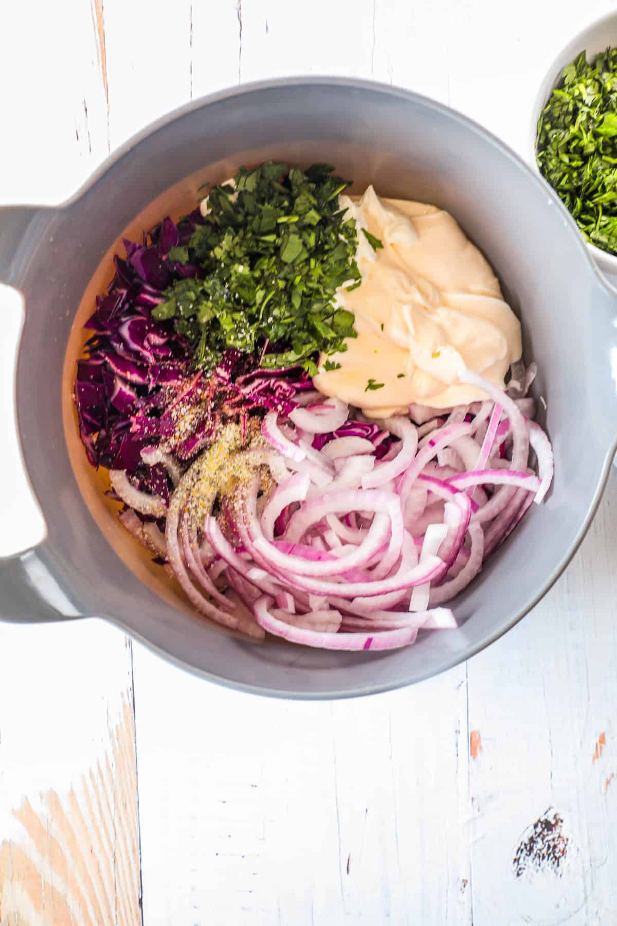 A mixing bowl containing shredded red cabbage, sliced red onions, chopped parsley, mayonnaise, and seasonings, prepared on a light wooden surface with additional chopped parsley in a small bowl nearby.