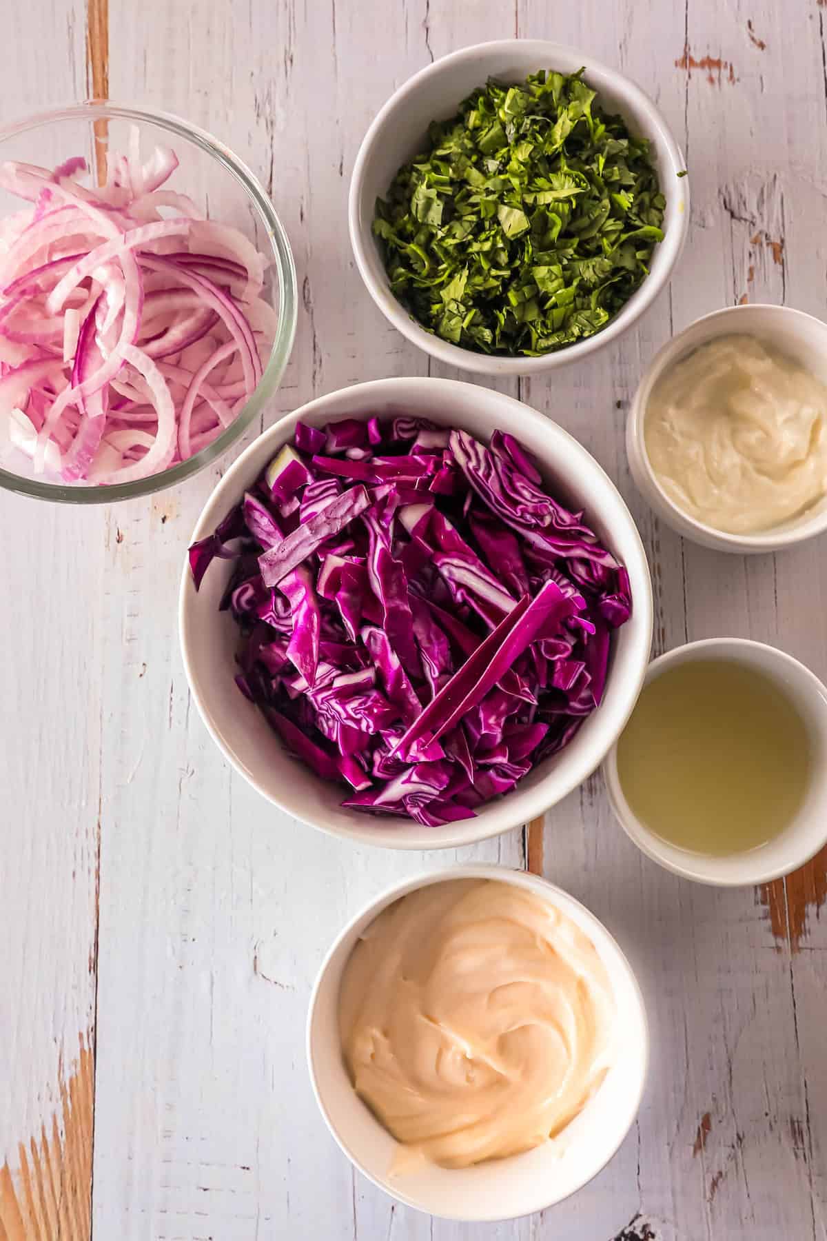 Top view of coleslaw ingredients arranged in bowls on a white wooden surface. Ingredients include red cabbage, red onion, parsley, mayonnaise, lemon juice, and a creamy dressing.