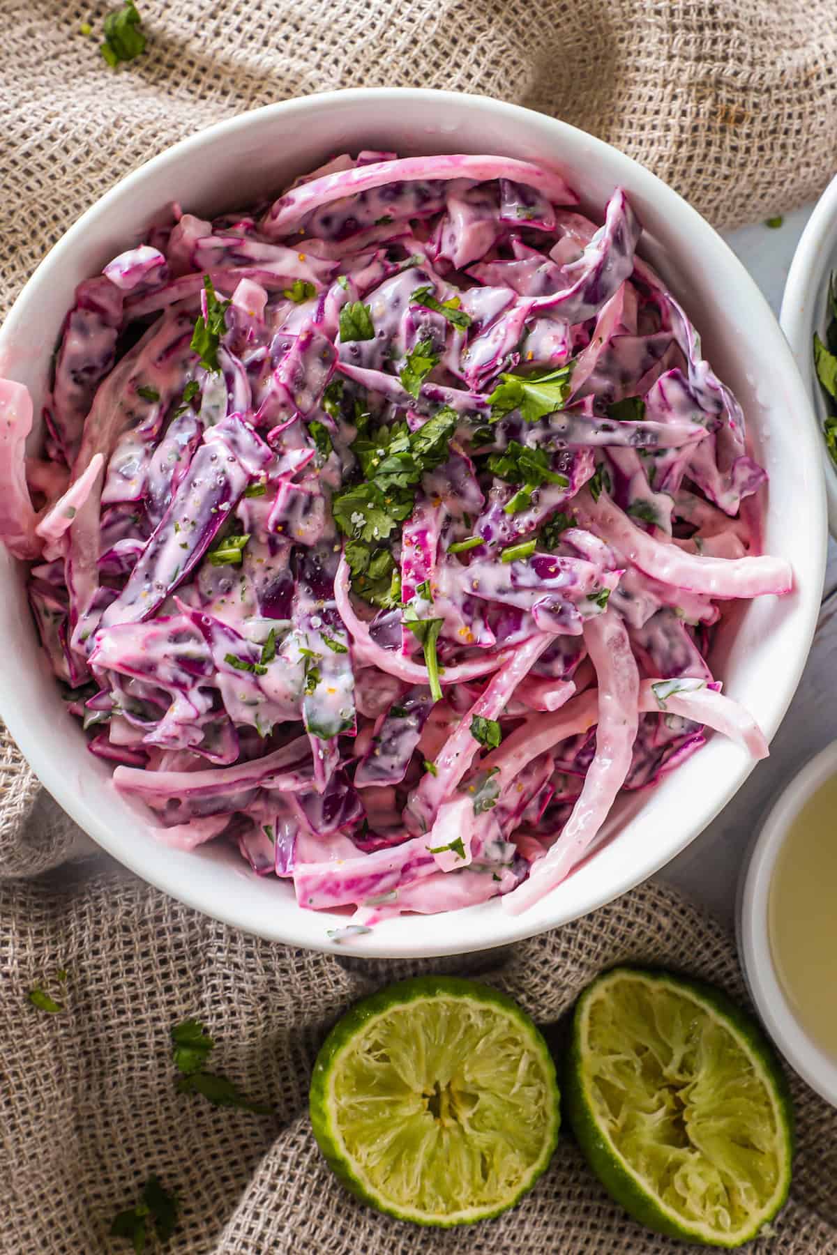 A bowl of creamy coleslaw made with purple and white cabbage, garnished with chopped cilantro. Two halved limes are placed beside the bowl on a textured beige surface.