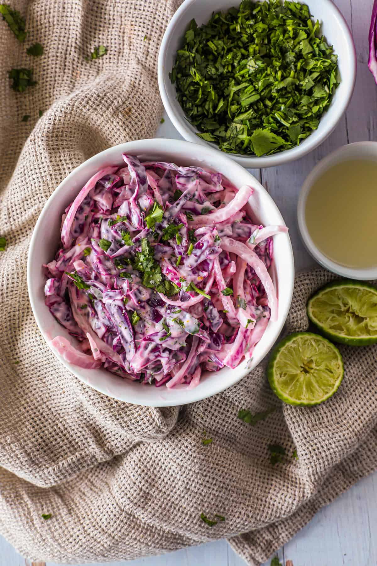 A bowl of coleslaw with red cabbage and green herbs on a cloth, accompanied by a bowl of chopped herbs, a cut lime, and a cup of light-colored liquid.