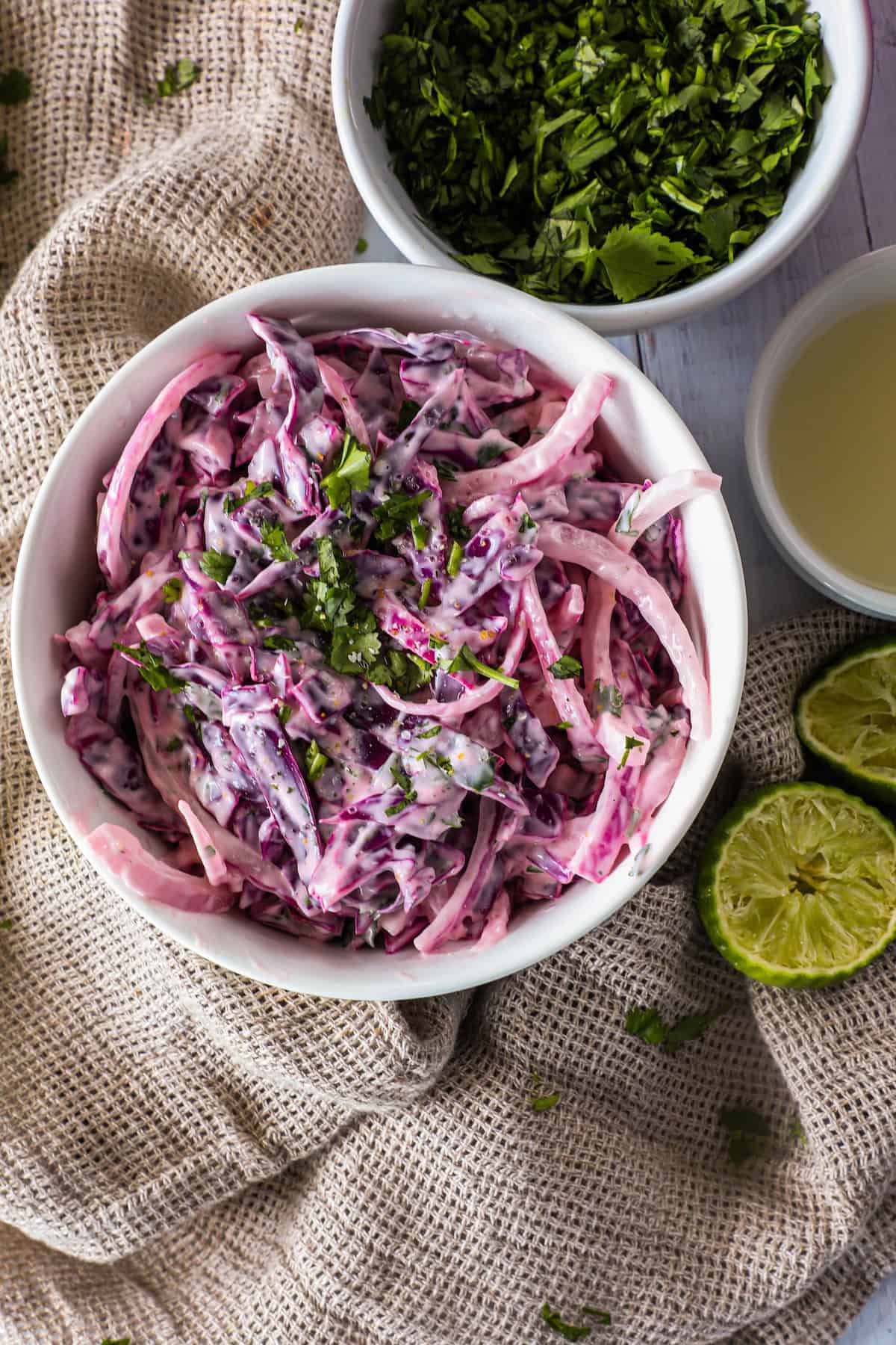 A bowl of creamy coleslaw with purple cabbage and onions, garnished with fresh herbs, placed on a textured fabric. Bowls of chopped herbs and lime juice, as well as halved limes, are nearby.