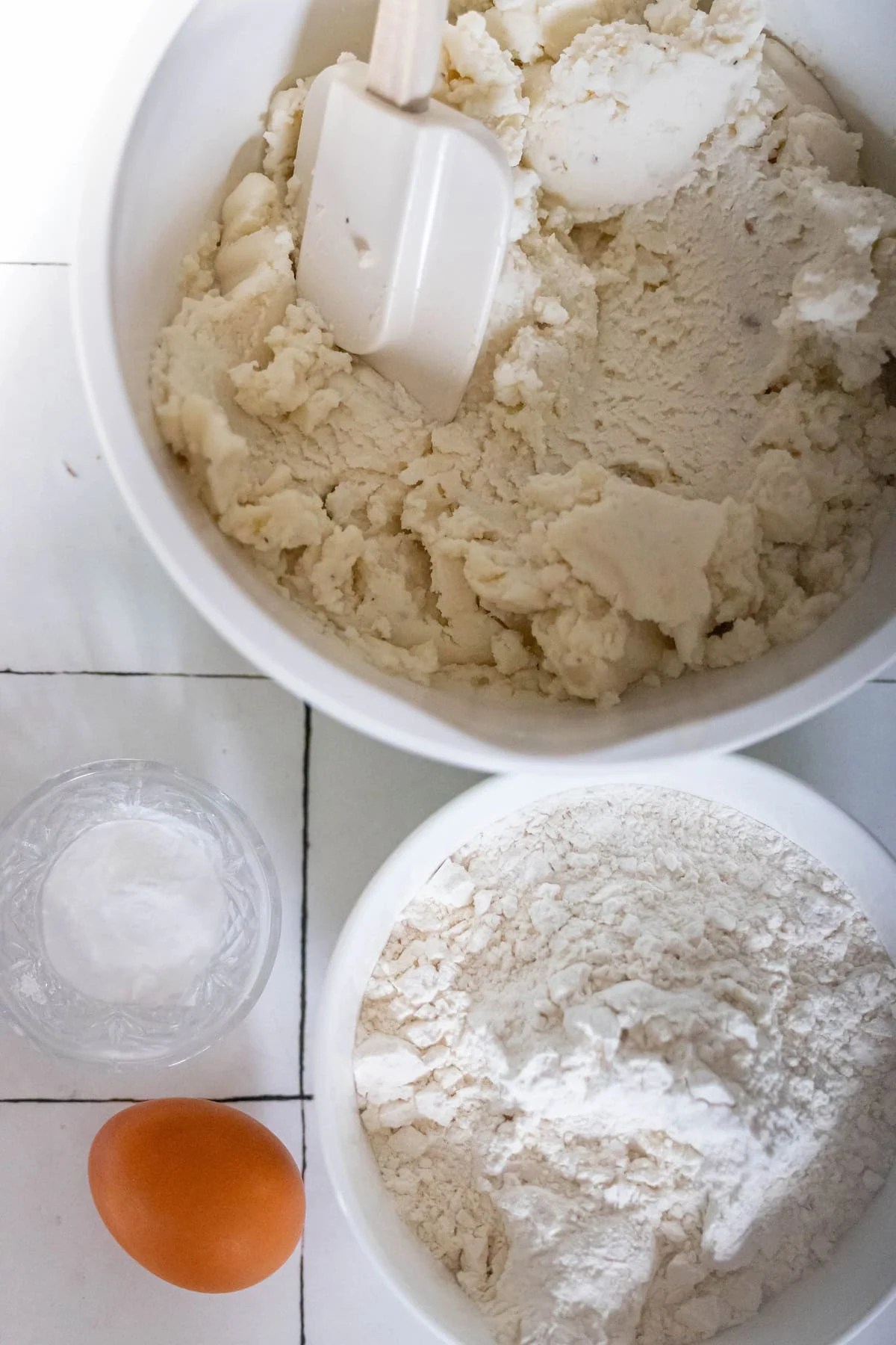 Bowls with dough, a spatula, flour, an egg, and a small glass of salt are arranged on a white tiled surface, ready for crafting the perfect Scottish Tattie Scones.