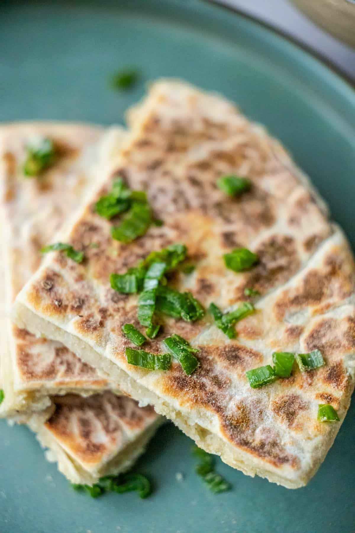 Sliced scallion pancakes on a green plate, reminiscent of Scottish tattie scones, topped with a sprinkle of chopped green onions.