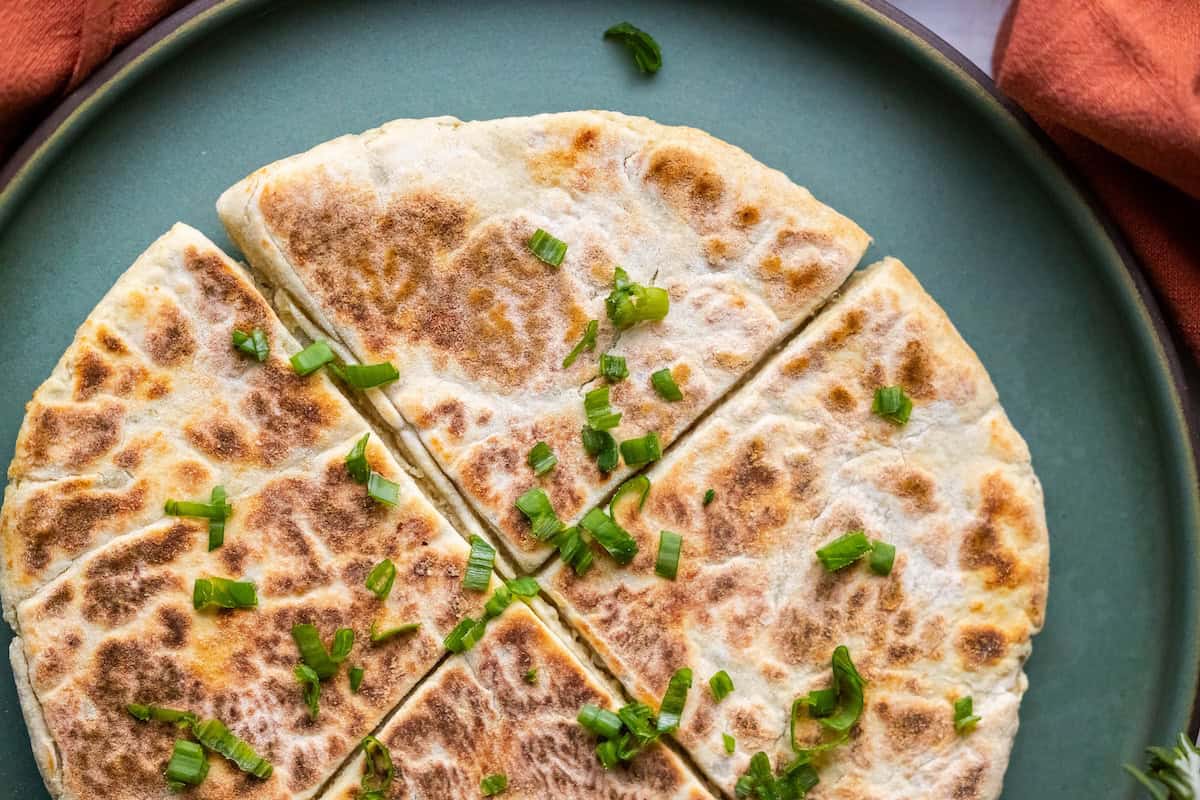 A sliced flatbread resembling Scottish tattie scones, topped with chopped green onions, rests on a green plate against an orange fabric background.