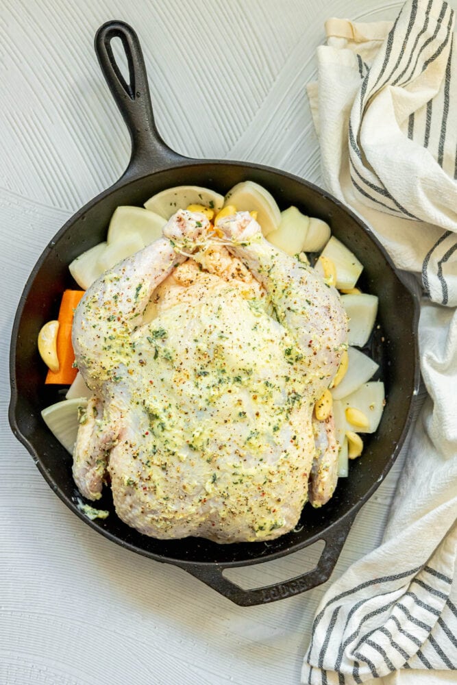 picture of a raw whole chicken with butter all over it on top of garlic, onions, and carrots in a cast iron skillet