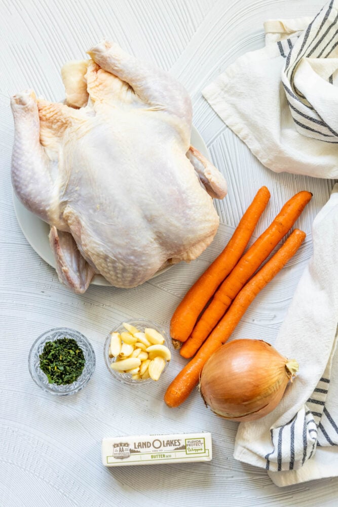 picture of a raw whole chicken, carrots, herbs, butter, garlic, and an onion on a table