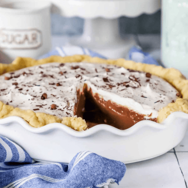 A freshly baked chocolate pie with a slice cut out, garnished with chocolate shavings, in a white pie dish on a blue-striped cloth.