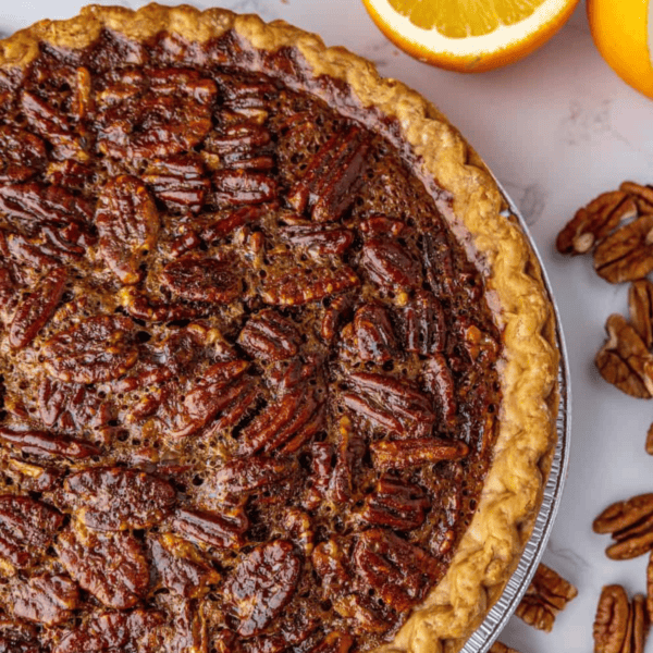 A flaky golden crust encases the rich pecan pie, perfectly displayed on a marble surface, enhanced by scattered pecans and the vibrant touch of half an orange.