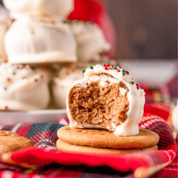 Close-up of a partially eaten white chocolate-covered gingerbread truffle with sprinkles on a gingham cloth. A stack of more gingerbread truffles is visible in the background.