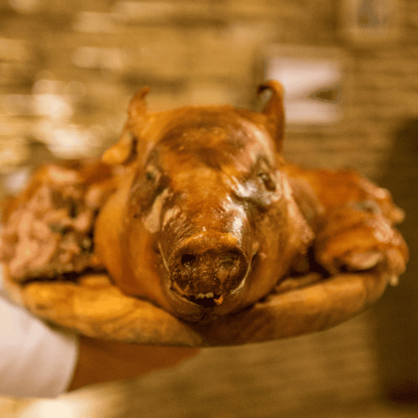 A roasted pig's head is presented on a wooden platter, emphasizing the pig's face and ears, reminiscent of a traditional Georgian Supra. The background is artfully blurred to highlight the centerpiece, evoking the rich traditions of a wine feast.