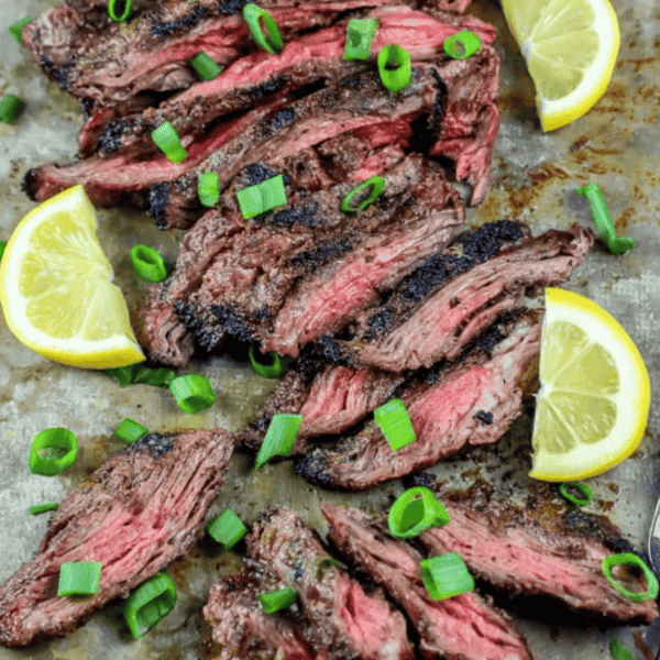 Sliced garlic grilled flank steak garnished with scallions and lemon wedges, all artfully arranged on a metal tray.