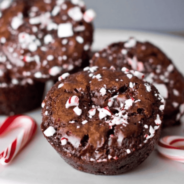 Peppermint brownies topped with crushed candy canes are stacked on a white plate, with a whole candy cane beside them. These delightful treats can be ready in just 30 minutes, combining rich chocolate with festive peppermint flair.