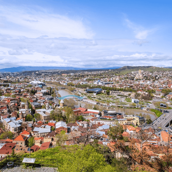 Aerial view of a city with a river flowing through it, numerous buildings, bridges, and a mix of natural landscapes and urban areas under a partly cloudy sky—a scene right out of a travel guide to Tbilisi.