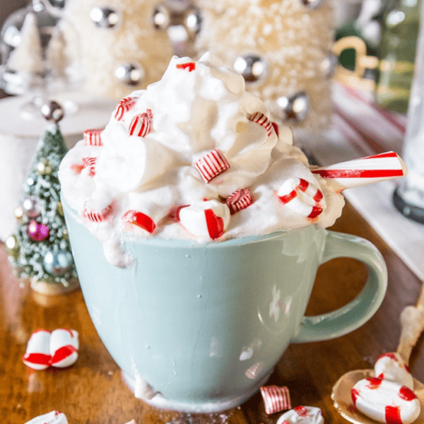 A light blue mug filled with whipped cream, topped with red and white peppermint candies and a candy cane stick, on a wooden surface with festive holiday decorations in the background, reminiscent of a cozy white cocoa peppermint latte.